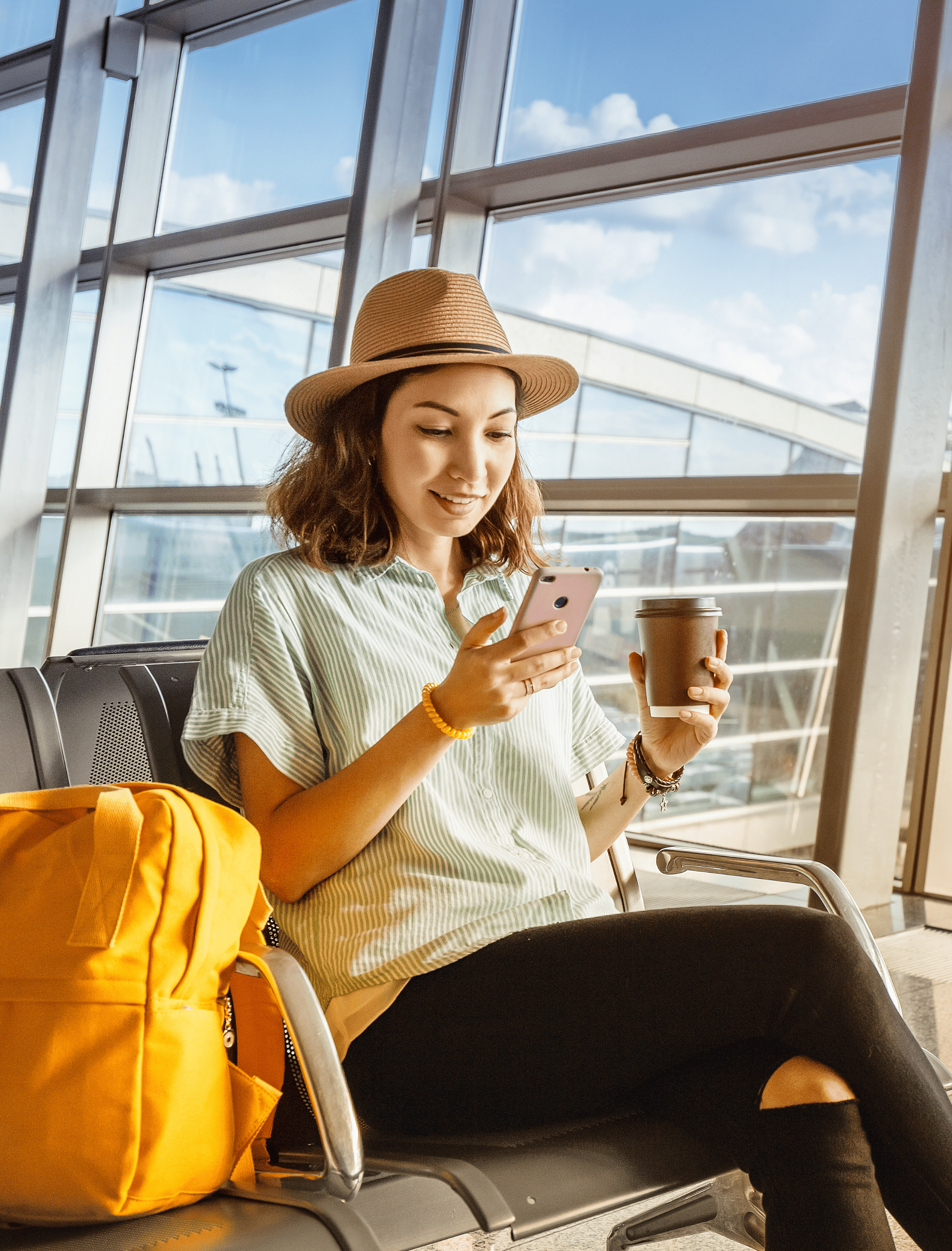 Young female traveller sitting at the airport on her phone with coffee in hand.